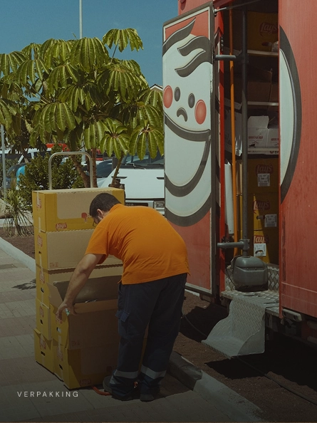 Een bezorger in een oranje t-shirt tilt kartonnen dozen van een steekwagen naast een geopende rode bestelwagen onder een boom