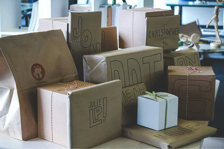 Een verzameling in bruin papier ingepakte cadeaudozen met handgeschreven namen zoals Christopher, Julie en Maude, samen met een papieren tas op tafel