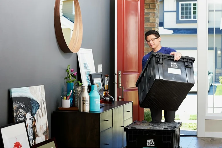 Een man met een bril draagt een grote zwarte plastic verhuisdoos van Hive Boxx door een open rode voordeur een modern ingericht huis binnen