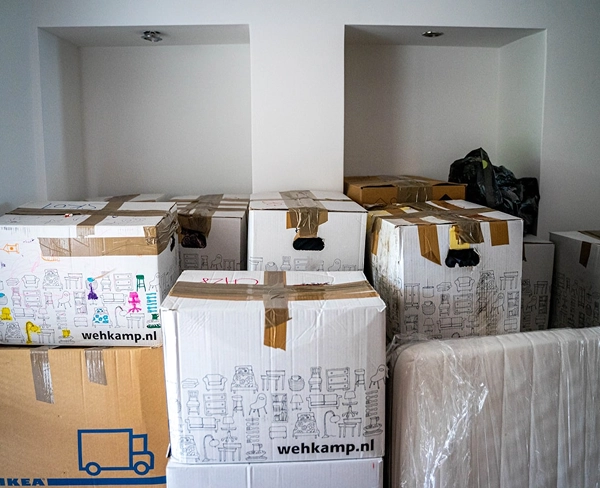 Een stapel kartonnen verhuisdozen met logo's van Wehkamp en Ikea die in een witte kamer met nissen staan opgesteld